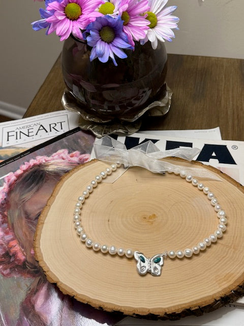 Pearl necklace with a butterfly clasp on a wooden surface, surrounded by flowers and a magazine.